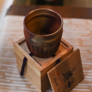 May include: A brown ceramic cup with a textured surface and gold accents sits inside a wooden box with a lid. The lid has Japanese characters written on it.