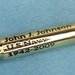 May include: A gold-colored bullet casing with black engraved text that reads "John J. Johnson U.S. Navy 1942-2009".