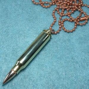 May include: A gold-colored bullet casing necklace with a copper-colored chain. The bullet casing is about 2 inches long and has a pointed tip. The chain is about 18 inches long and has a lobster clasp.