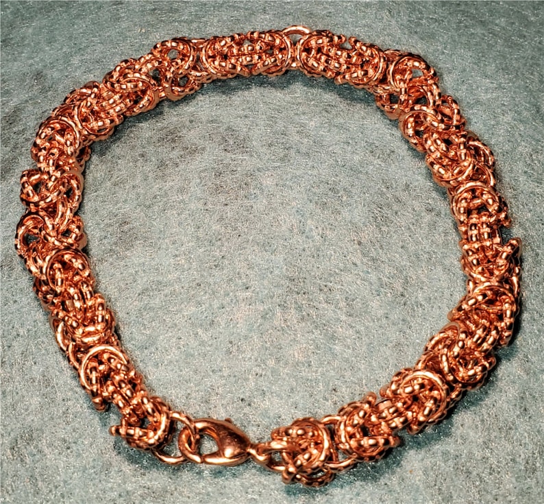 May include: A copper-colored bracelet with a woven design. The bracelet is made of small, interconnected rings that create a intricate pattern.