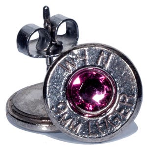 May include: A pair of silver bullet earrings with a pink rhinestone in the center. The bullet casings are marked "9mm Luger" and "Win".