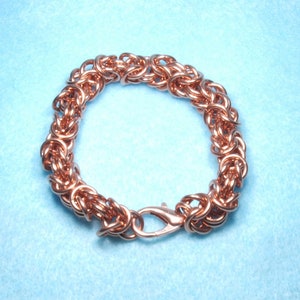 May include: A copper-colored Byzantine chainmail bracelet with a lobster clasp. The bracelet is made of small, interlocking rings, creating a textured, woven appearance. The jewelry is displayed on a light blue surface.