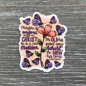 May include: A sticker with a watercolor background featuring a pink flower with green leaves and purple butterflies. The sticker includes the text "Therefore, if anyone is in Christ he is a new creation. The old has passed away; behold, the new has come. 2 Corinthians 5:17"