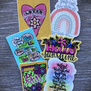 May include: Assortment of colorful stickers with inspirational quotes and designs. Stickers feature phrases like "Loved beyond measure", "I know the plans I have for you", "Be Still", "Hello Sunshine", and "The Lord bless you and keep you".