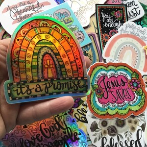 May include: A collection of colorful stickers with religious themes. The stickers feature rainbows, crosses, and inspirational messages like "It's a promise", "Jesus Saves", and "Love God and People".