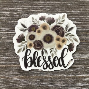 May include: A white sticker with a floral design in shades of brown, beige, and purple. The word "blessed" is written in black cursive lettering below the flowers.