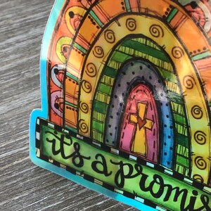May include: A colorful sticker with a rainbow arch and a cross in the center. The sticker has a green banner with the text "it's a promise" in black cursive writing.