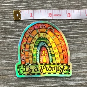May include: A colorful rainbow sticker with a cross in the center and the text "it's a promise" below. The sticker is iridescent and has a holographic finish.