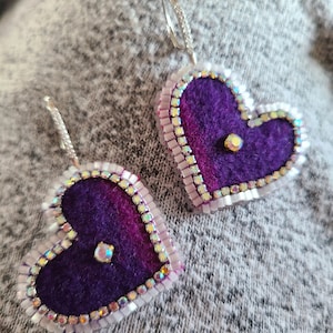 Pode incluir: Um par de brincos de coração roxo com strass iridescentes ao redor das bordas. Os brincos são feitos de feltro e têm um gancho prateado.