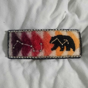 May include: A rectangular hair clip featuring a felt background with a gradient of purple, red, and orange hues. The design includes a beaded black bear silhouette and a constellation pattern made of small clear crystals, bordered by black beads.