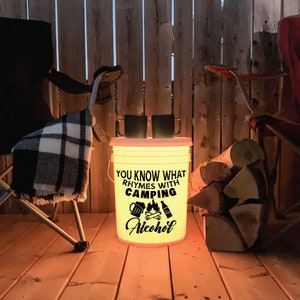 May include: A white bucket with a black graphic that reads "You know what rhymes with camping? Alcohol." The bucket is lit from within, casting a warm glow. Two chairs and a pile of firewood are in the background.