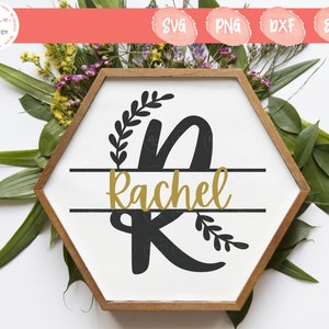 May include: A black and white sign with a gold name on it. The sign is in the shape of a hexagon and has a wooden frame. The name on the sign is "Rachel" with a decorative "R" and a wreath of leaves around it.