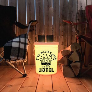 May include: A white bucket with a black design that says "Five Billion Star Hotel" with a camper and stars. The bucket is lit from inside with a warm yellow light. Two black mugs are on top of the bucket. The bucket is on a wooden deck with two folding chairs and a pile of firewood.