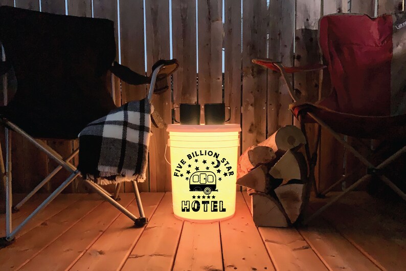 May include: A white bucket with a black design that says "Five Billion Star Hotel" with a camper and stars. The bucket is lit from inside with a warm yellow light. Two black mugs are on top of the bucket. The bucket is on a wooden deck with two folding chairs and a pile of firewood.