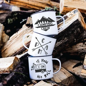 May include: Three white enamel camping mugs stacked on top of each other. The top mug has the text "Camping Crew" with a mountain range graphic. The middle mug has the text "Camp" with crossed arrows. The bottom mug has the text "This is how we roll" with a camper van graphic.
