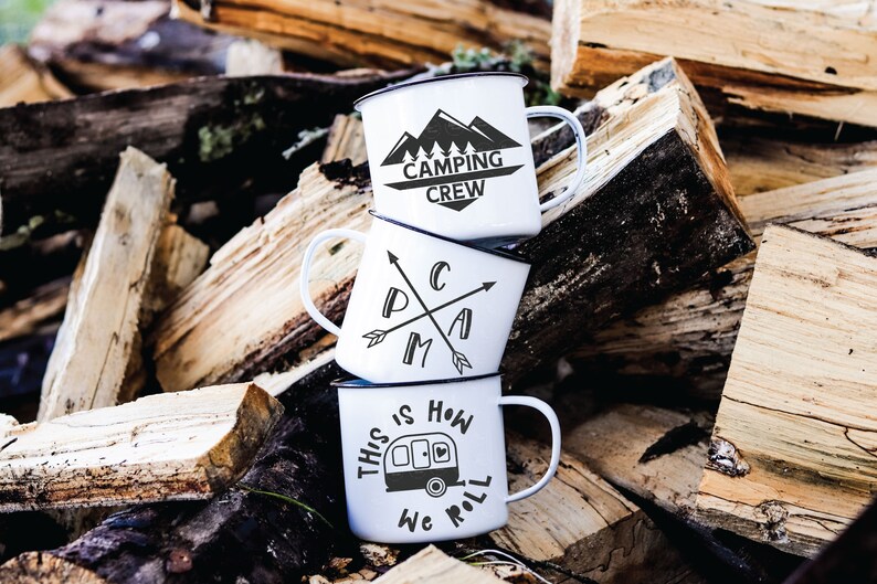 May include: Three white enamel camping mugs stacked on top of each other. The top mug has the text "Camping Crew" with a mountain range graphic. The middle mug has the text "Camp" with crossed arrows. The bottom mug has the text "This is how we roll" with a camper van graphic.