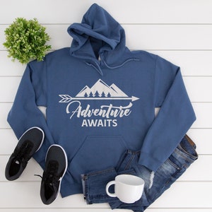May include: A blue hooded sweatshirt with a white graphic of mountains, trees, and an arrow with the text "Adventure Awaits". The sweatshirt is laid out on a white wooden surface with a pair of black sneakers, blue jeans, and a white mug.