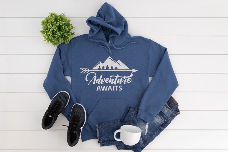 May include: A blue hooded sweatshirt with a white graphic of mountains, trees, and an arrow with the text "Adventure Awaits". The sweatshirt is laid out on a white wooden surface with a pair of black sneakers, blue jeans, and a white mug.