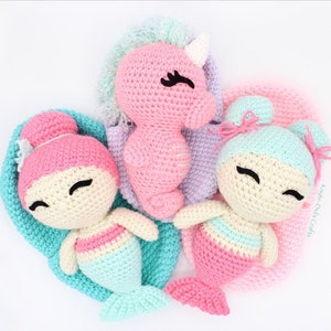 Może przedstawiać: Trzy pluszowe zabawki wykonane na szydełku: różowy jednorożec, różowo-niebieska syrena i niebiesko-różowa syrena. Zabawki uśmiechają się i mają czarne oczy. Jednorożec ma biały róg i niebieską grzywę. Syreny mają różowo-niebieskie ogony.