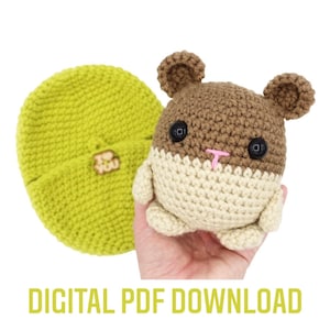May include: Crochet pattern for a brown and cream hamster amigurumi toy. The pattern includes instructions for making the hamster's body, head, ears, and nose. The pattern also includes instructions for making a small green crocheted hat.