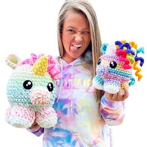 May include: A woman wearing a tie-dye hoodie holds two crocheted rainbow unicorn plushies. The larger unicorn is white with rainbow stripes and a yellow horn. The smaller unicorn is white with rainbow stripes and a purple, pink, and yellow horn.