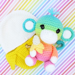 May include: A crocheted monkey toy holding a banana. The monkey is green, pink, and yellow. The banana is cream and yellow. The monkey has a white face with black eyes. The monkey is sitting on a rainbow striped background.