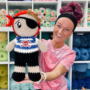 May include: A crocheted pirate doll with a black eye patch, red hair, and a blue and white striped shirt with a skull and crossbones design. The doll is being held by a person with pink hair and a pink shirt with a map design.