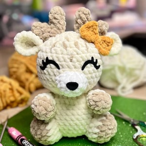 May include: A handmade crochet deer with a cream-colored body, brown antlers, and an orange bow. The deer has black eyelashes and a black nose. The deer is sitting on a green surface. A pink crochet hook and scissors are also visible.