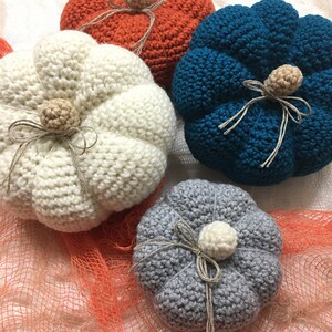 May include: Four crocheted pumpkins in different colors: white, orange, teal, and gray. Each pumpkin has a stem and a bow made of twine.