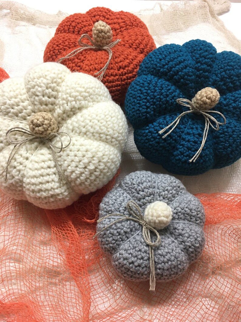 May include: Four crocheted pumpkins in different colors: white, orange, teal, and gray. Each pumpkin has a stem and a bow made of twine.
