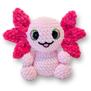 Puede incluir: Un juguete de peluche axolotl de crochet rosa con ojos grandes e iridiscentes y una expresión feliz. El axolotl tiene branquias rosadas de crochet.