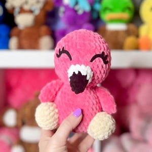 May include: A pink crocheted flamingo toy with white and black facial features. The toy is being held in a hand with purple nail polish.