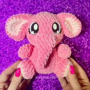 May include: A pink crocheted elephant plush toy with white accents on the ears and a sparkly pink nose. The toy has large, black eyes with white pupils and a pink, crocheted body.