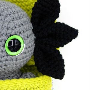 May include: A gray crocheted amigurumi toy with a green eye and black crocheted ears. The toy is sitting in a yellow crocheted basket.