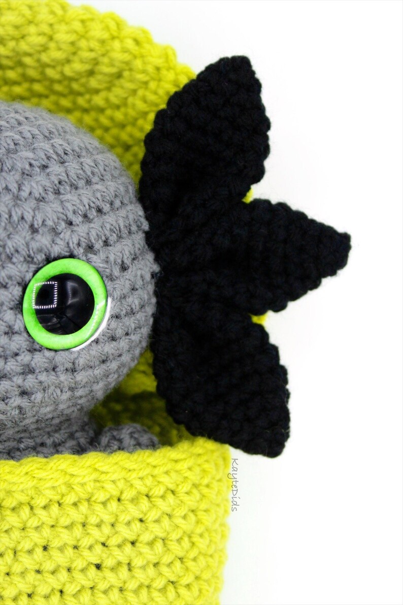 May include: A gray crocheted amigurumi toy with a green eye and black crocheted ears. The toy is sitting in a yellow crocheted basket.