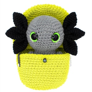 May include: A gray crocheted axolotl plush toy with black fins and green eyes, sitting inside a yellow crocheted pouch.