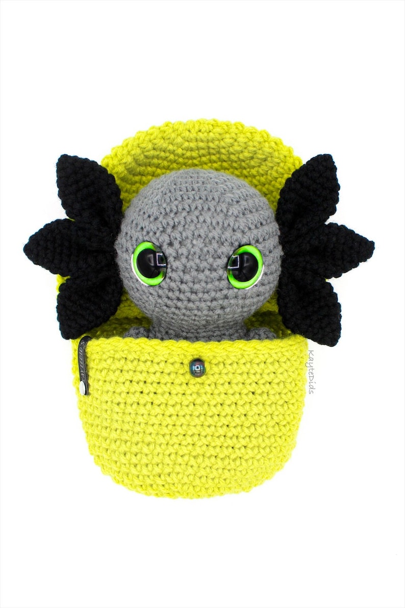 May include: A gray crocheted axolotl plush toy with black fins and green eyes, sitting inside a yellow crocheted pouch.