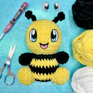 May include: A crocheted bee plush toy with black and yellow stripes, big eyes, and a smiling mouth. The bee is sitting on a blue surface with yarn balls in the background.