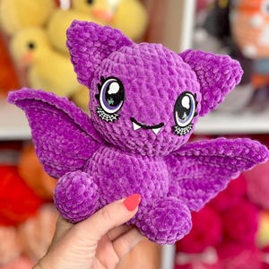 May include: A plush, purple bat with large, expressive eyes and detailed wings. The handmade bat has a smiling mouth and small fangs. The bat is held in front of a shelf with other plush toys.