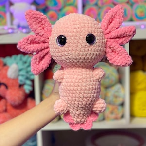 May include: A pink crocheted axolotl plush toy with large black eyes. The toy is being held by a hand in front of a shelf with colorful yarn.