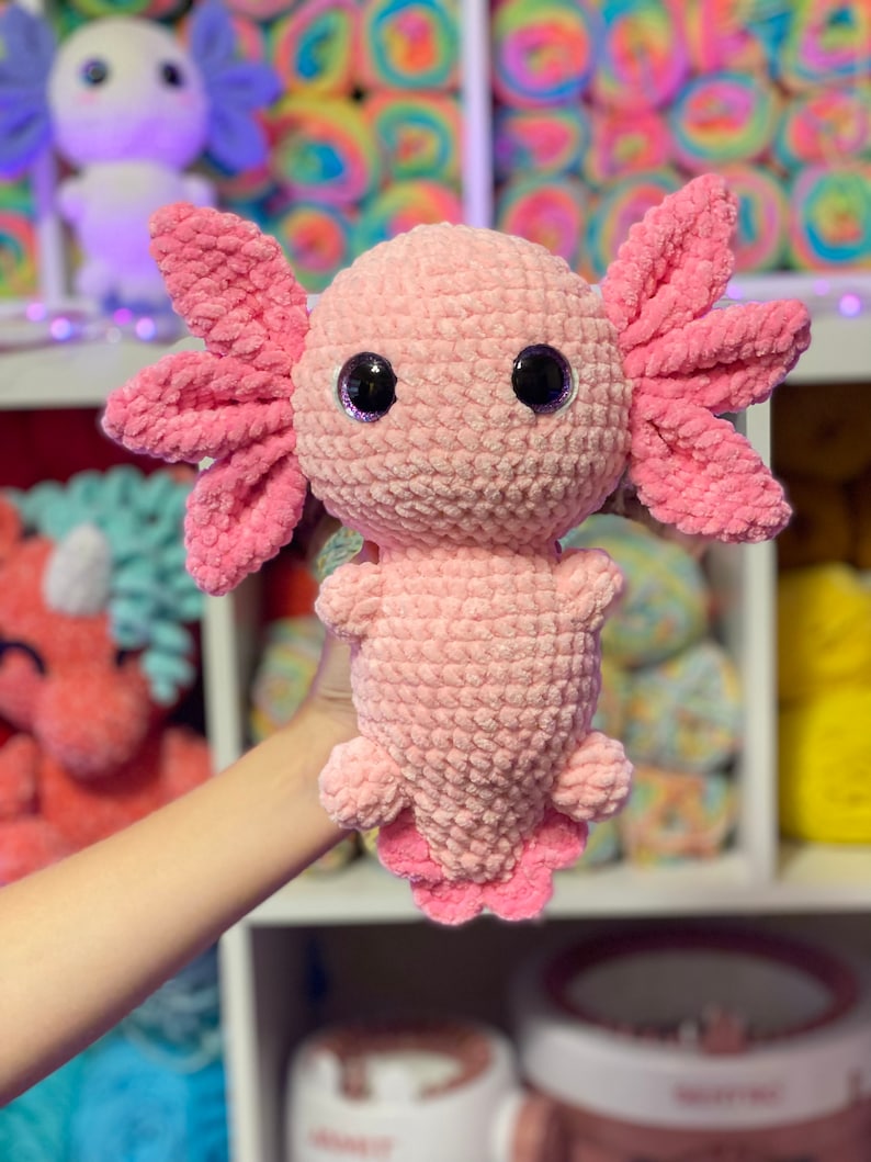 May include: A pink crocheted axolotl plush toy with large black eyes. The toy is being held by a hand in front of a shelf with colorful yarn.