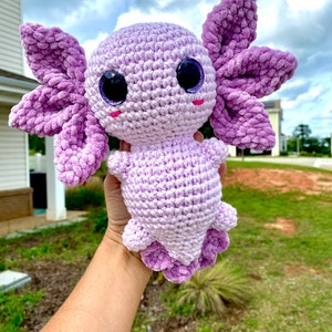 May include: A crocheted axolotl plush toy with purple and white yarn. The toy has large, glittery eyes and a pink blush on its cheeks.
