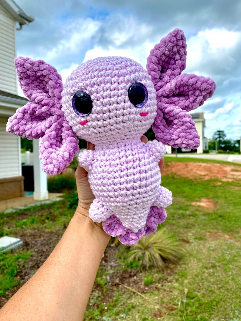 May include: A crocheted axolotl plush toy with purple and white yarn. The toy has large, glittery eyes and a pink blush on its cheeks.