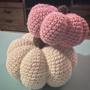May include: Two crocheted pumpkins, one white and one pink, stacked on top of each other. The white pumpkin is larger and has a more rounded shape, while the pink pumpkin is smaller and has a more pointed shape.