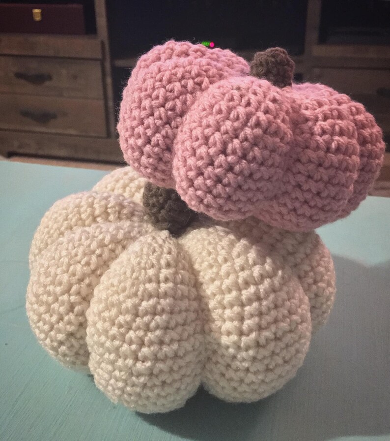 May include: Two crocheted pumpkins, one white and one pink, stacked on top of each other. The white pumpkin is larger and has a more rounded shape, while the pink pumpkin is smaller and has a more pointed shape.