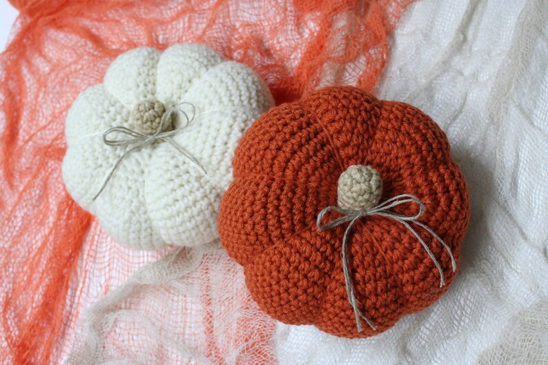 May include: Two crocheted pumpkins, one white and one orange, with brown twine bows and a small crocheted stem. The pumpkins are sitting on a white and orange mesh fabric.