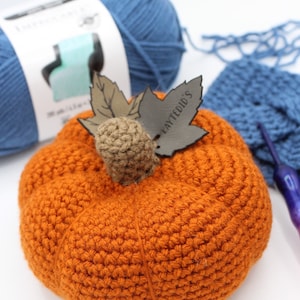 Könnte beinhalten: Ein gehäkelter orangefarbener Kürbis mit einem braunen Stiel und einem Blatt mit dem Text "Laytedid's". Der Kürbis liegt auf einer weißen Oberfläche mit blauem Garn und einer Häkelnadel.