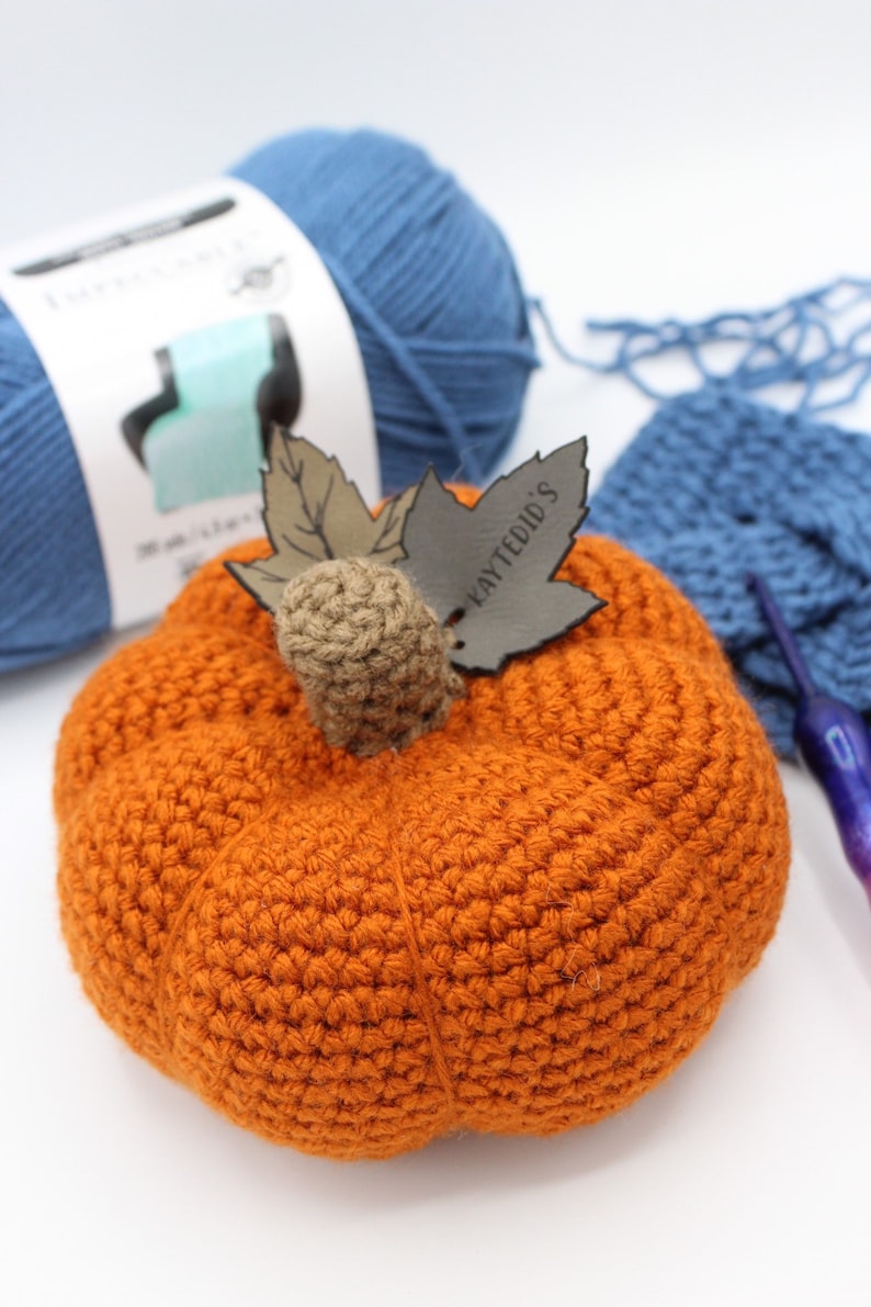 May include: A crocheted orange pumpkin with a brown stem and a leaf with the text "Laytedid's" on it. The pumpkin is sitting on a white surface with blue yarn and a crochet hook.