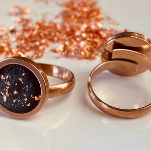 Könnte beinhalten: Zwei goldene Ringe mit einer schwarzen und goldenen Einlage. Der Ring links hat eine runde Einlage mit goldenen Flocken auf schwarzem Hintergrund. Der Ring rechts ist ein schlichtes Band.