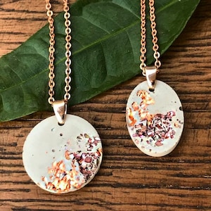 Könnte beinhalten: Zwei Halsketten mit kupfernen Ketten und runden und ovalen Anhängern aus weißem Beton mit eingebetteten schimmernden rosa und kupferfarbenen Flocken.